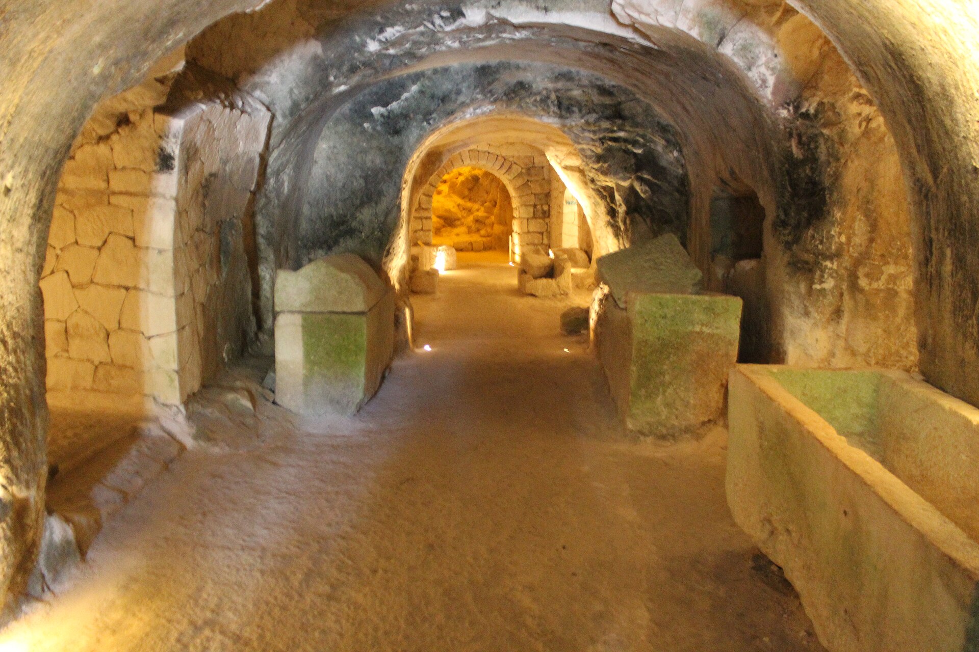 Corredor abobadado escavado na rocha, iluminado, com sarcófagos de pedra dos dois lados.