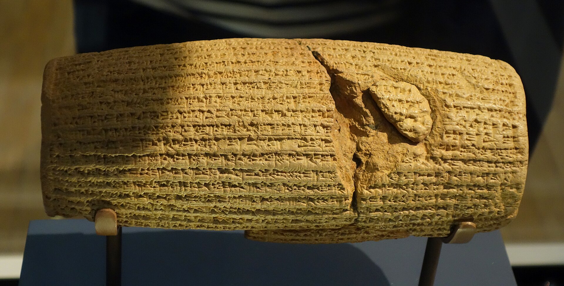 Cilindro de argila coberto de escrita cuneiforme, numa vitrine de museu.